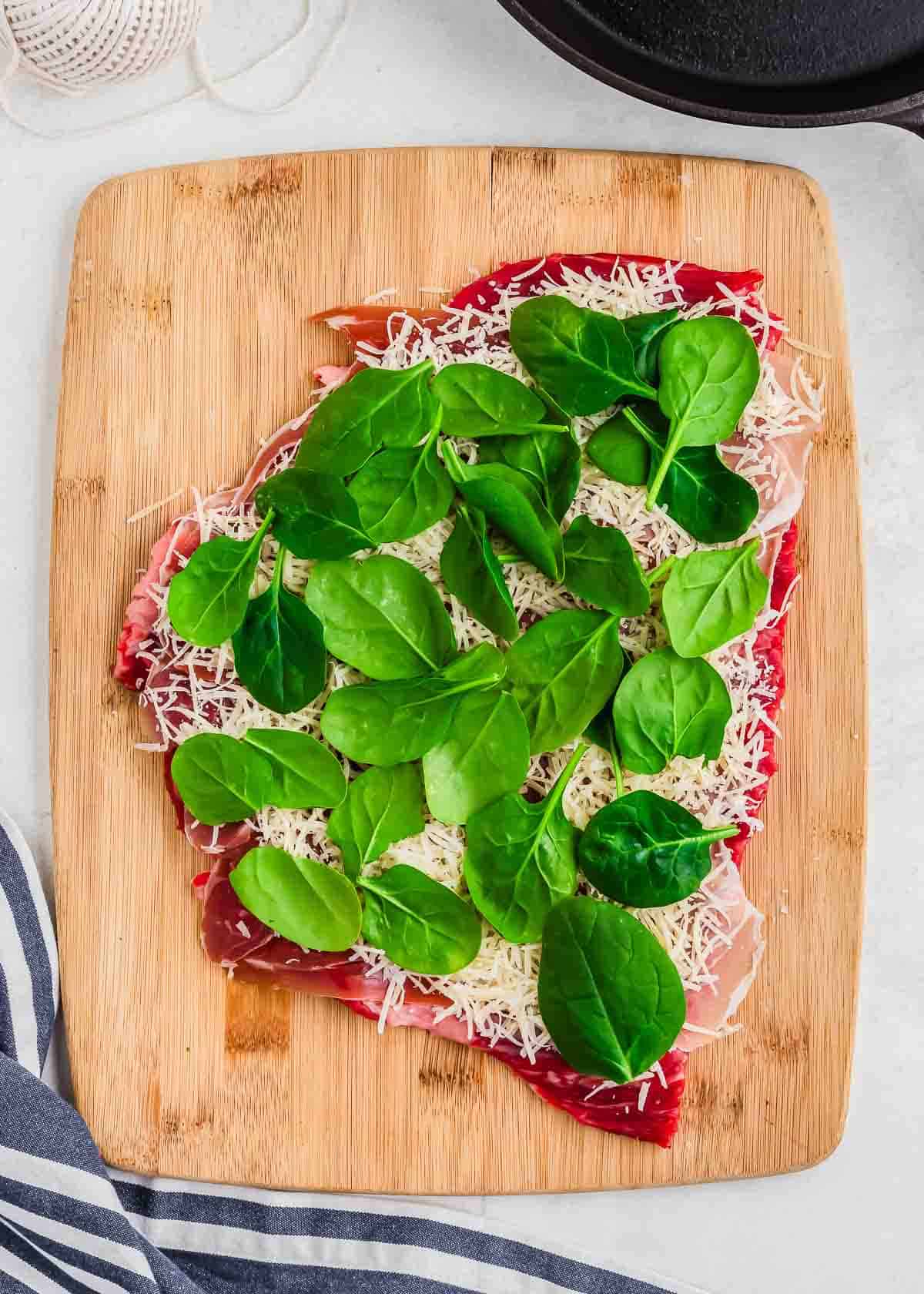 Thinly pounded flank steak on a cutting board topped with pesto, prosciutto, parmesan and baby spinach.
