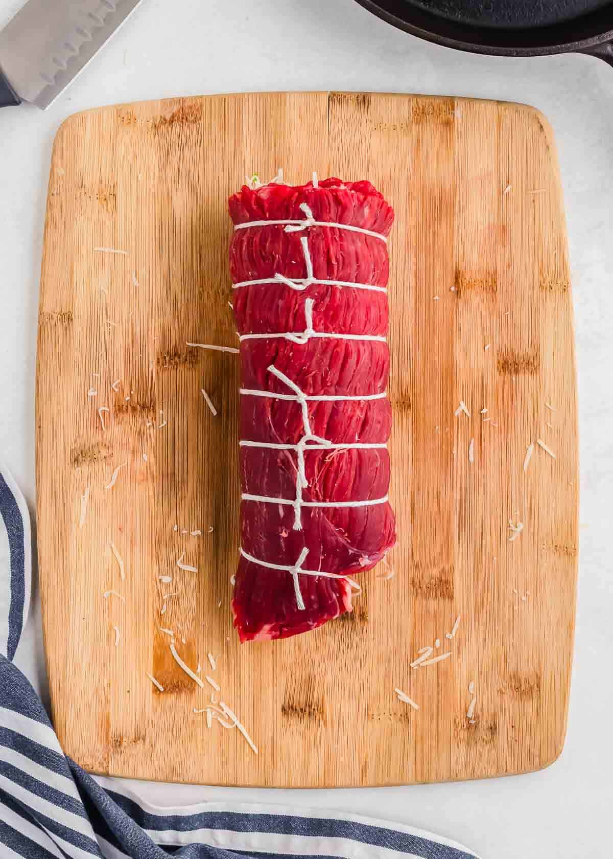 Raw flank steak rolled and tied with butcher's twine on a wooden cutting board.