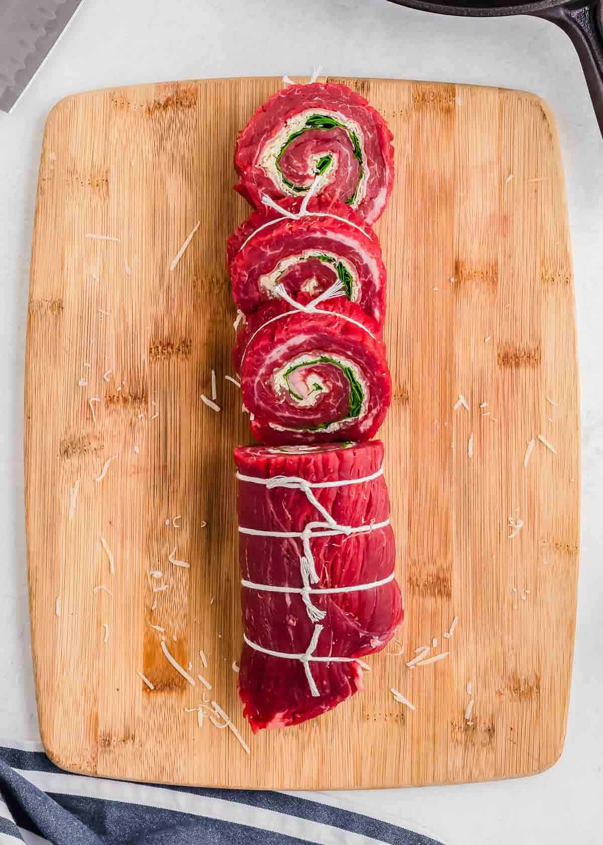 Raw pinwheel flank steaks with spinach and cheese filling on a wooden cutting board.