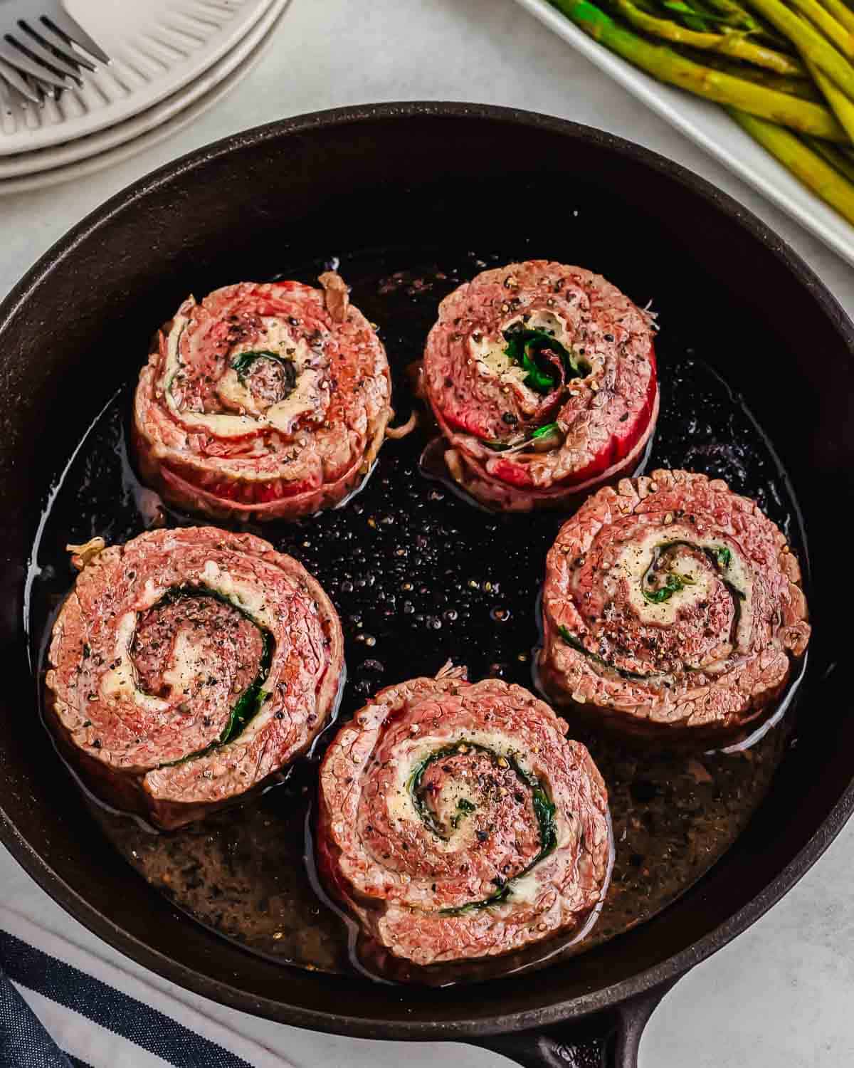 Five pinwheel stuffed steaks cooking in a cast iron skillet.