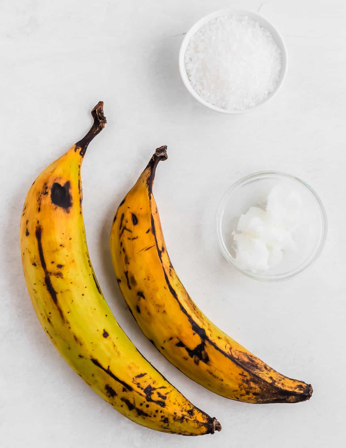 Two ripe plantains with black spots next to small bowls of salt and solidified coconut oil on a light surface.