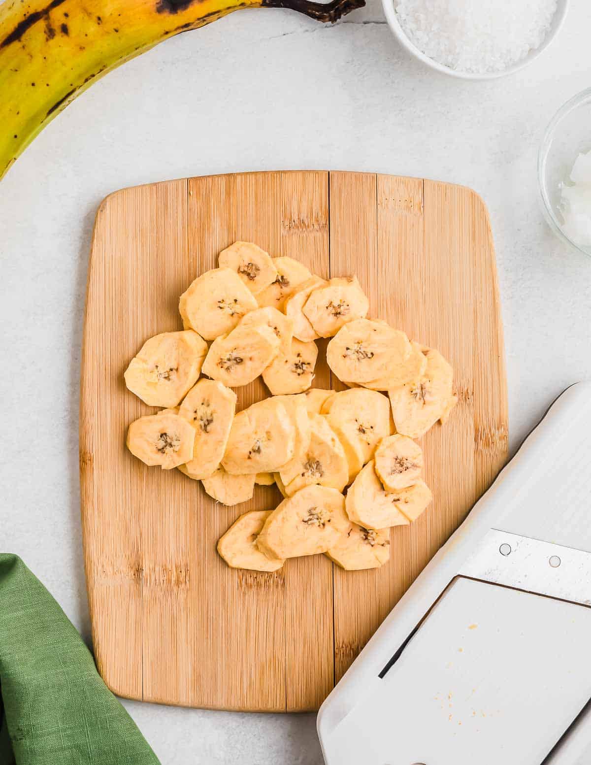 Sliced plantains on a wooden cutting board next to a knife and a sprinkling of coarse salt.