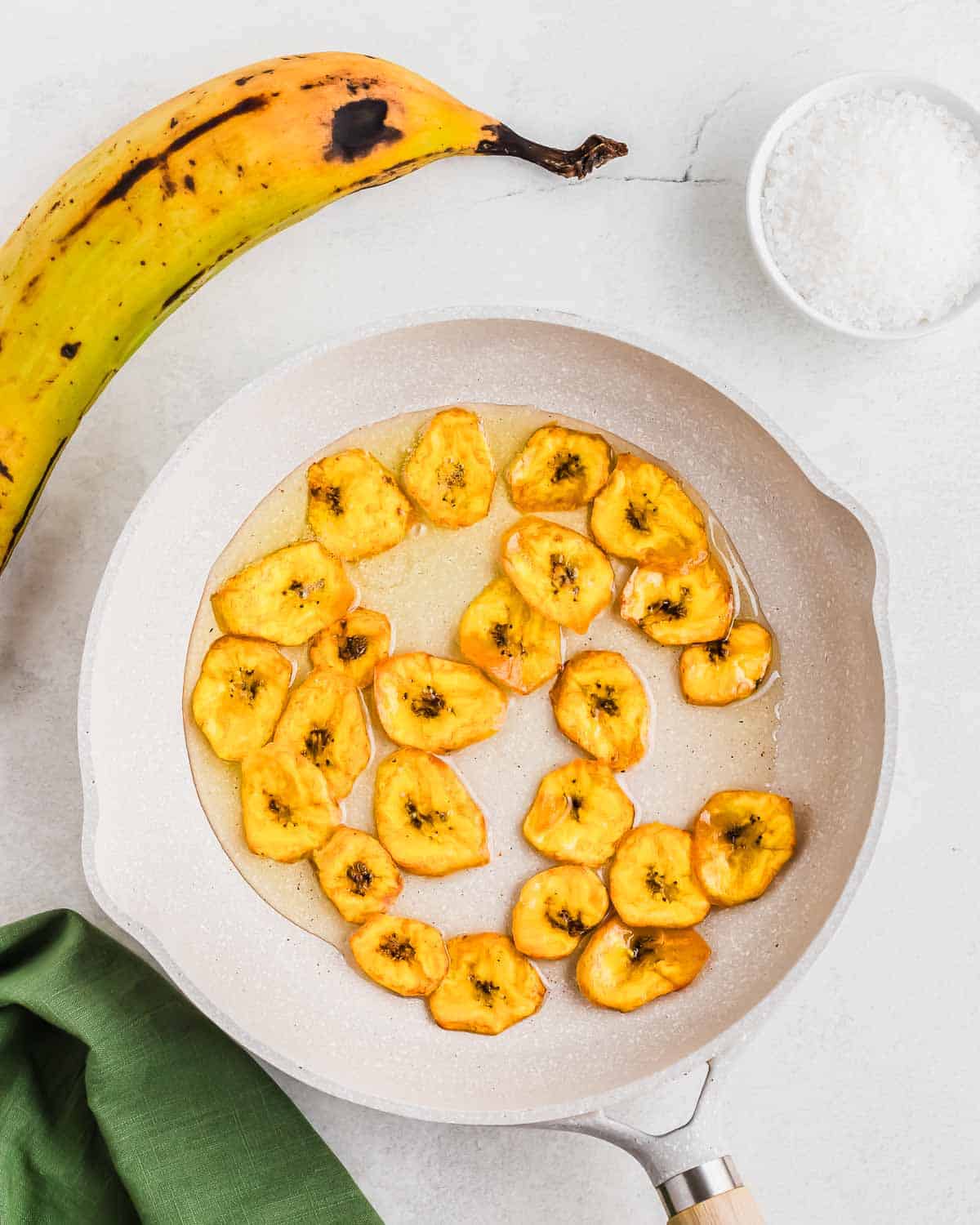 Sliced plantains being pan-fried with a pinch of salt to the side.