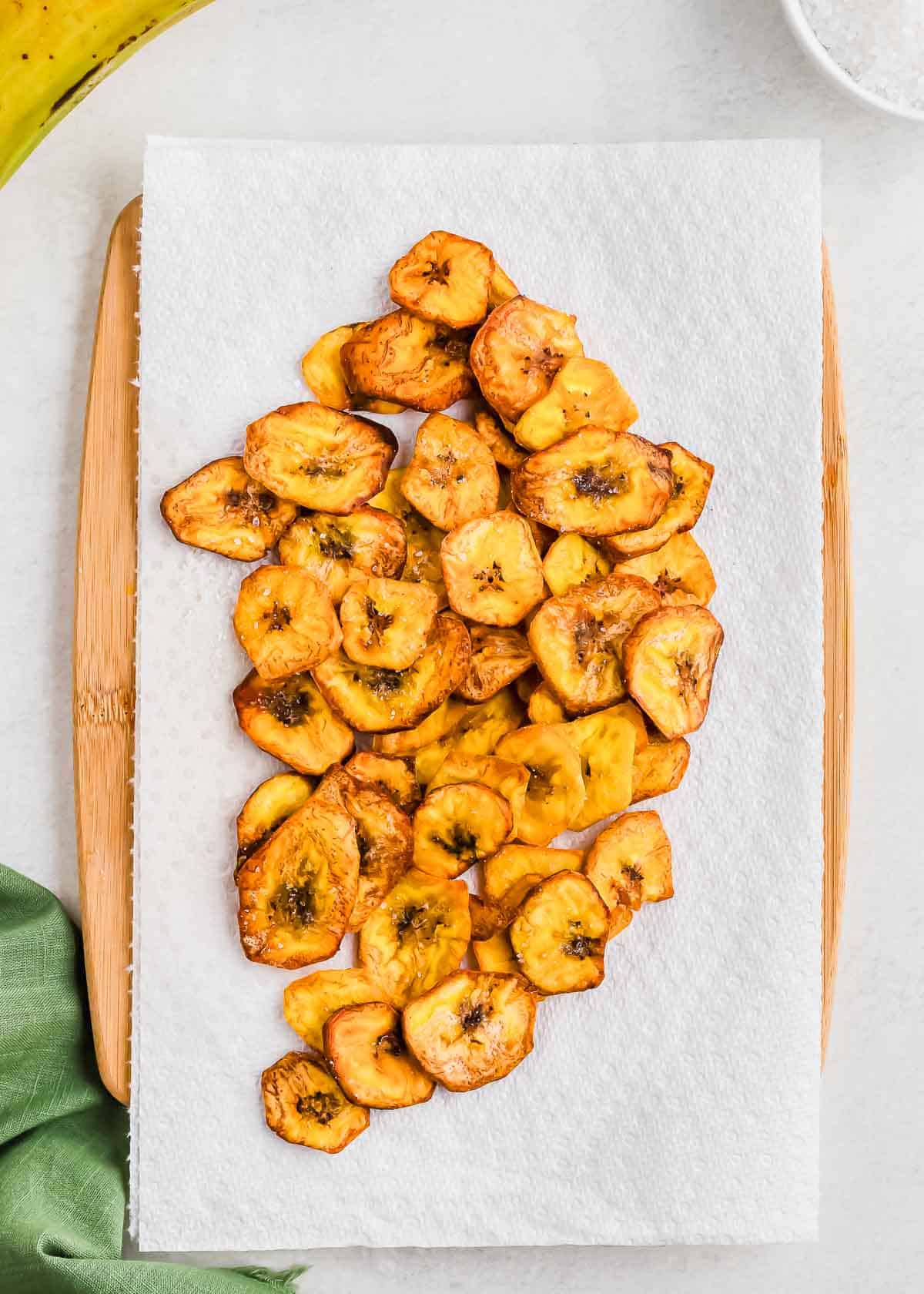 Fried plantains arranged on a paper towel beside a green towel and a wooden spatula.