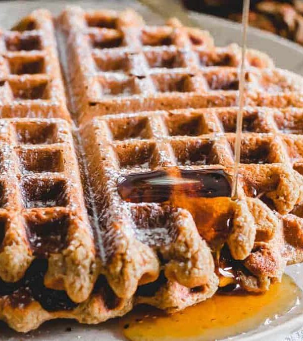 A freshly baked waffle on a plate with syrup being poured over it, surrounded by holiday decorations, including pine cones and red ornaments.
