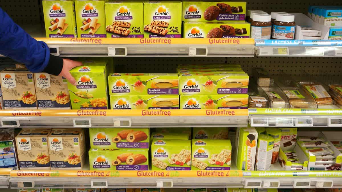 A person's hand selecting a box from a supermarket shelf stocked with gluten-free products, including cookies and snacks.