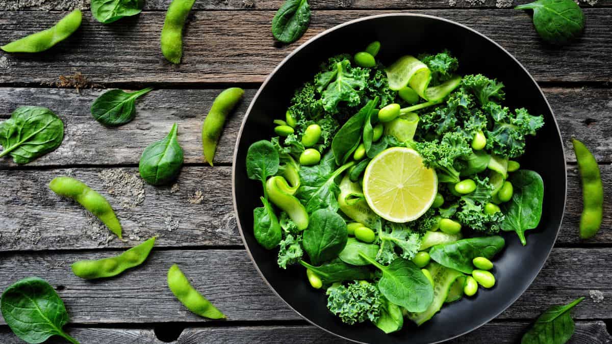 A vibrant green salad with kale, spinach, edamame, and a slice of lime served in a black bowl on a rustic wooden table.