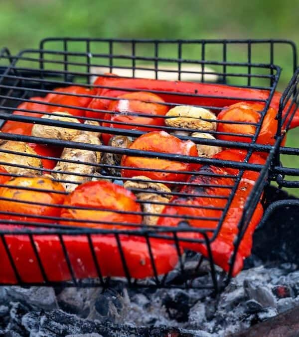 Grilled vegetables, including red peppers and mushrooms, cooking on a charcoal grill outdoors.