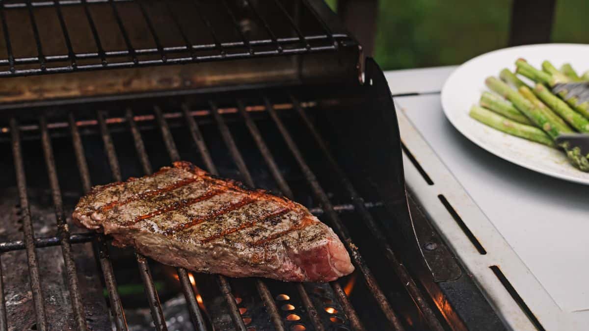 A steak with grill marks cooking on a barbecue next to a plate of grilled asparagus.