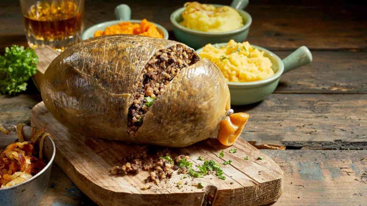 Scottish haggis on a wooden board surrounded by side dishes of mashed potatoes, turnips, and a glass of whisky.