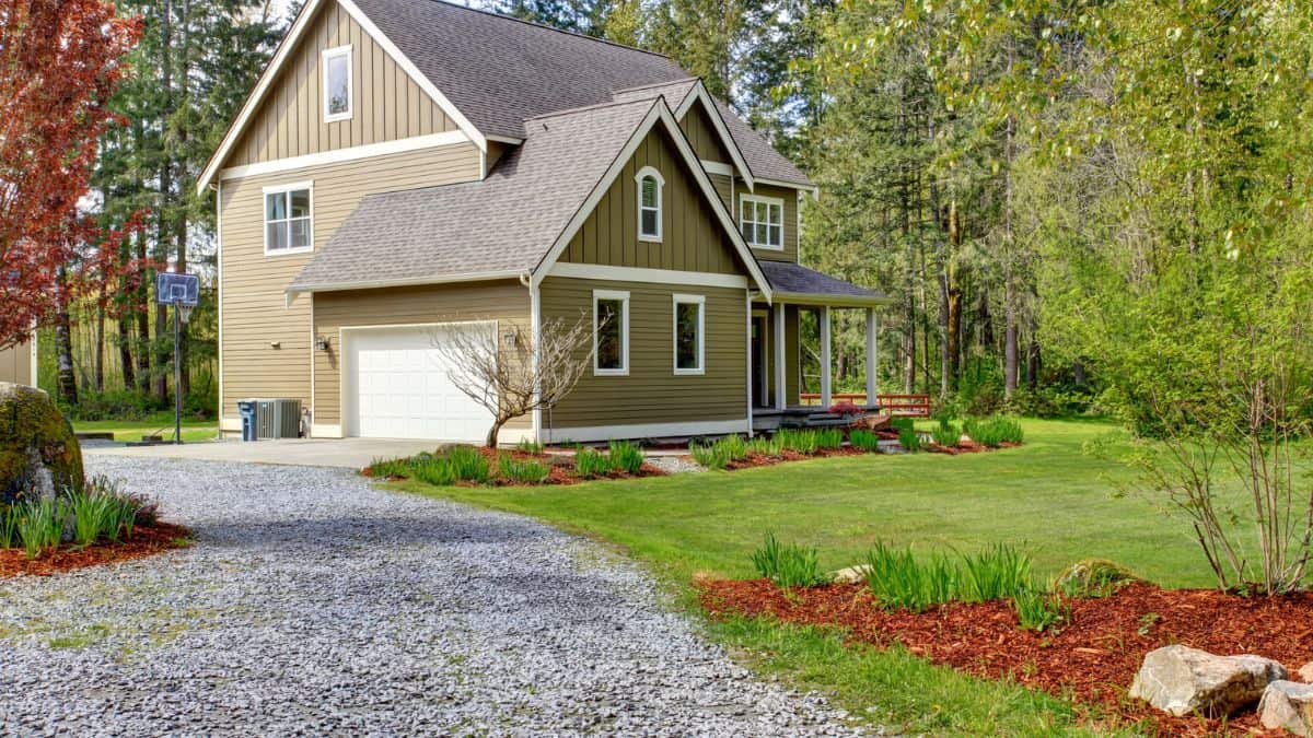 Two-story beige house with a double garage and a driveway, surrounded by lush greenery and a well-maintained lawn, under a clear sky.