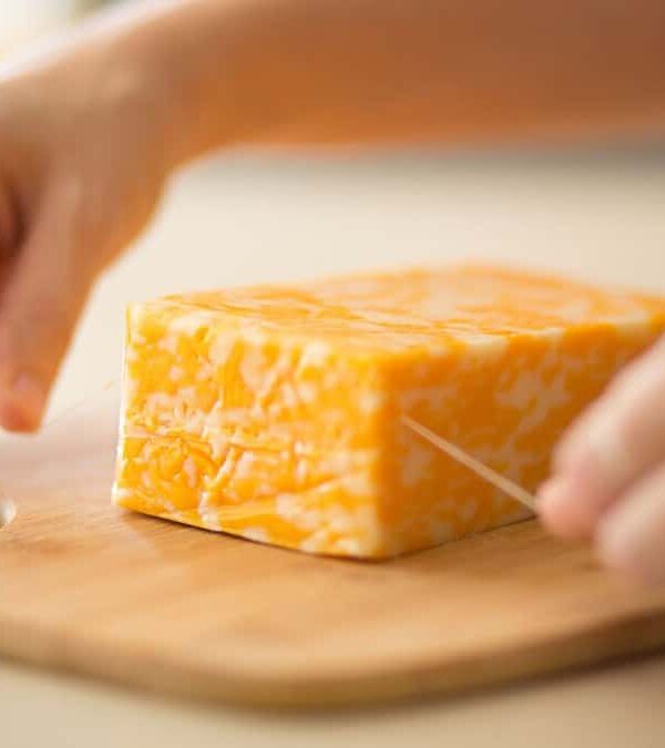 A person slicing a block of marbled cheddar cheese on a wooden cutting board.