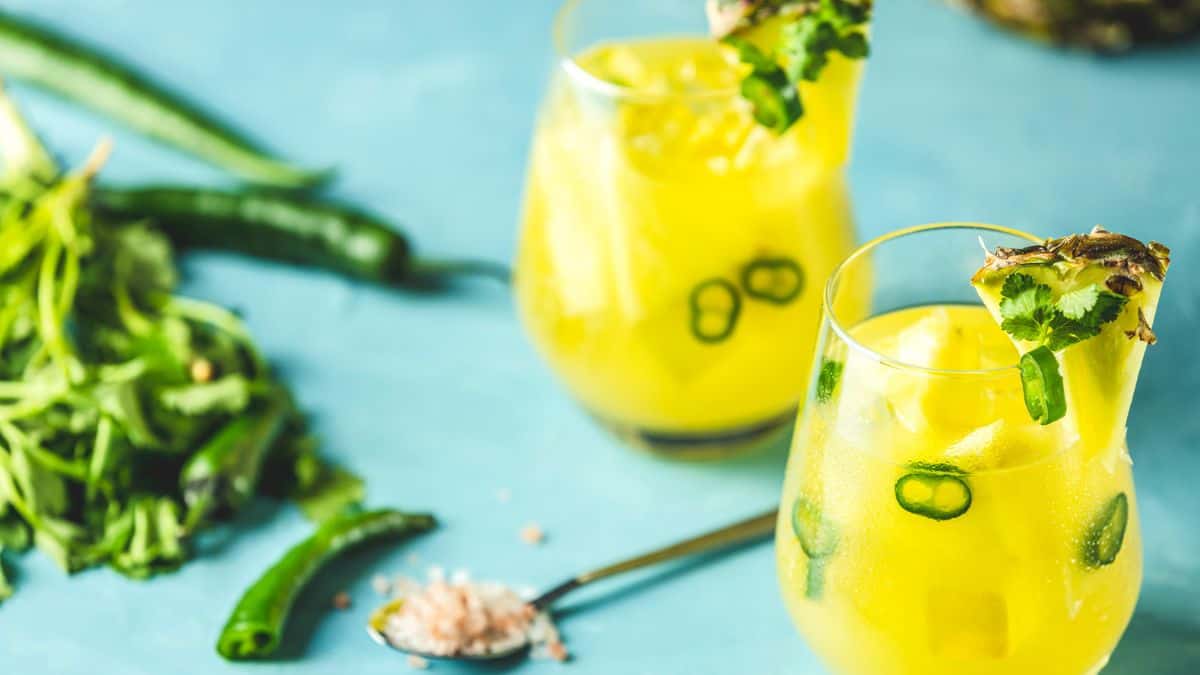 Two glasses of pineapple and jalapeño-infused beverages garnished with cilantro on a blue surface, accompanied by fresh ingredients.