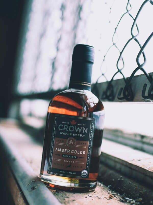 A bottle of crown maple syrup, amber grade, placed on a wooden surface near a window with a chain-link barrier.