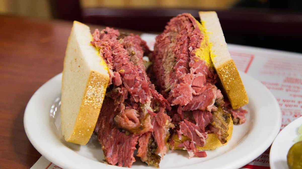 A thick pastrami sandwich on rye bread served on a white plate with pickles on the side.