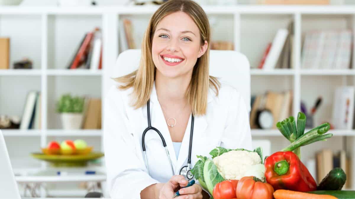 A female nutritionist with a stethoscope around her neck smiling at the camera, surrounded by fresh vegetables on her desk.