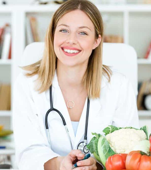 A female nutritionist with a stethoscope around her neck smiling at the camera, surrounded by fresh vegetables on her desk.