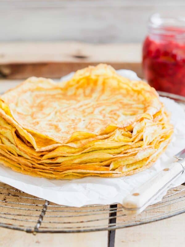 A stack of thin, golden crepes on a white plate beside a jar of red berry compote and a knife on a rustic wooden table.