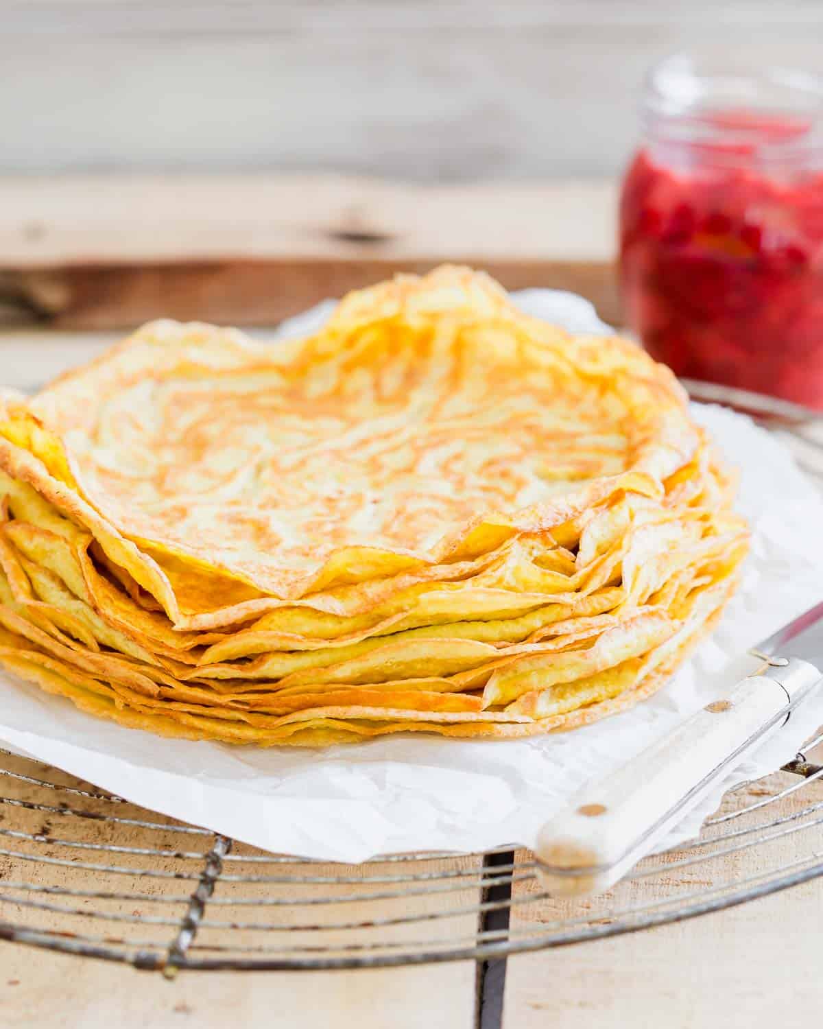 A stack of thin, golden crepes on a white plate beside a jar of red berry compote and a knife on a rustic wooden table.