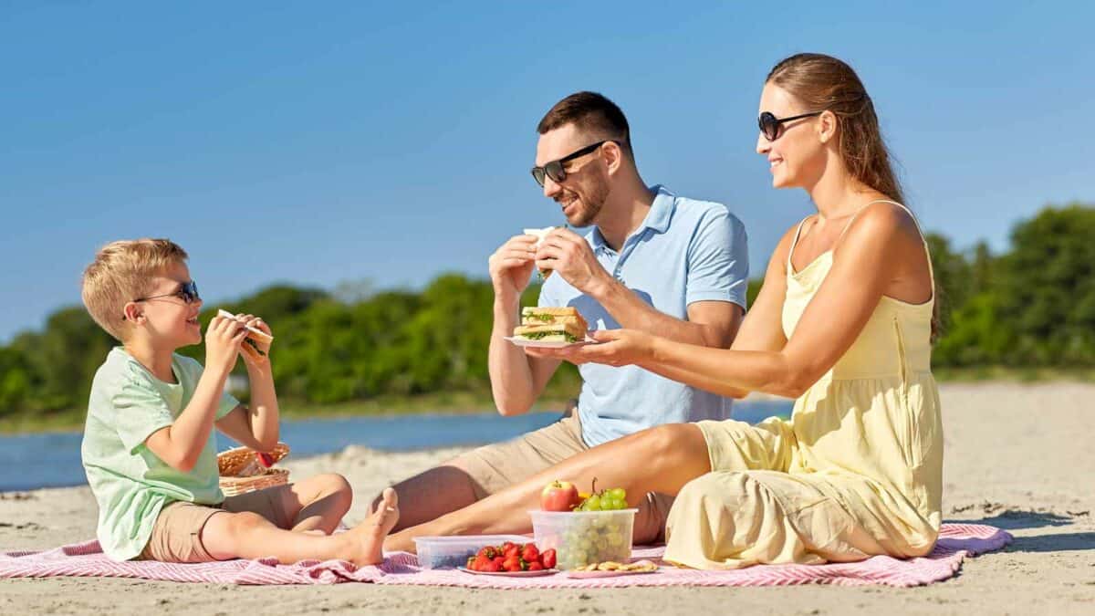 A family enjoying a picnic on a sunny beach, with a man, woman, and child eating sandwiches and fruits on a pink blanket.