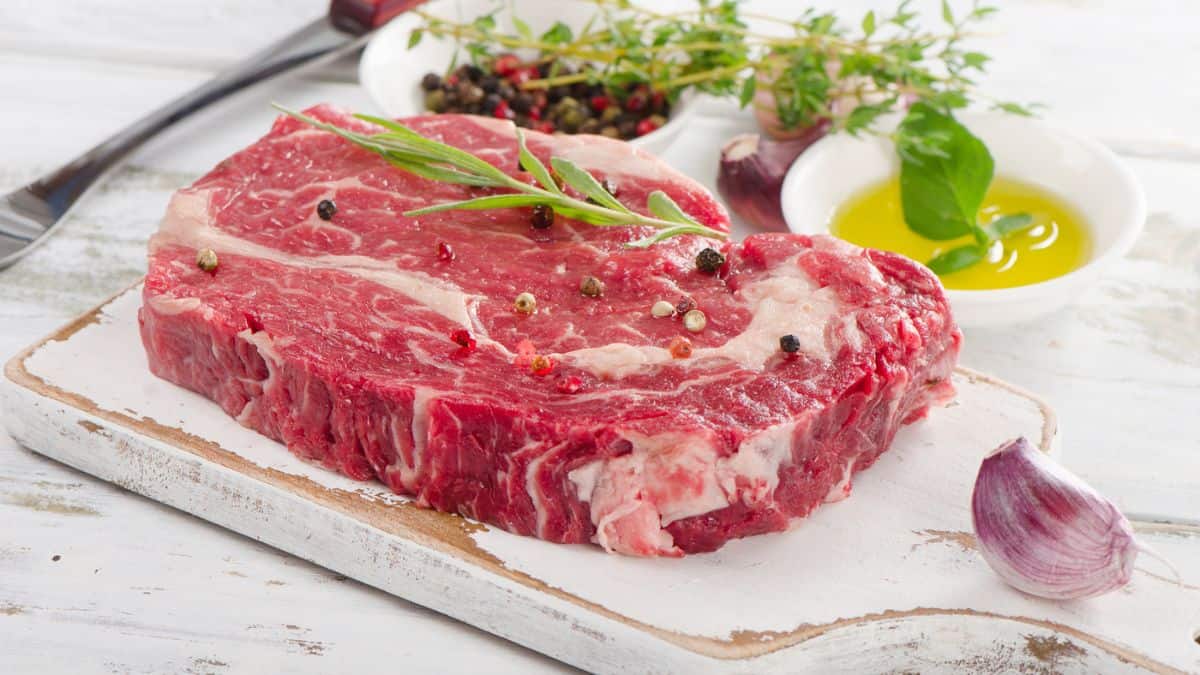 Raw steak seasoned with spices on a wooden board, accompanied by olive oil, garlic, and fresh herbs.