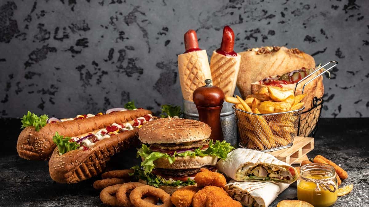 Assorted fast food items including burgers, hot dogs, wraps, onion rings, and fries on a wooden table with condiments.