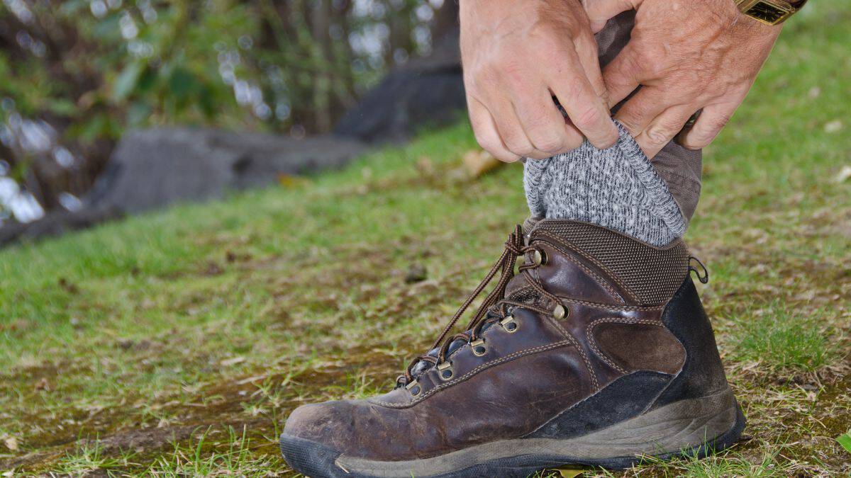 A person adjusting their gray sock while wearing a brown hiking boot outdoors.