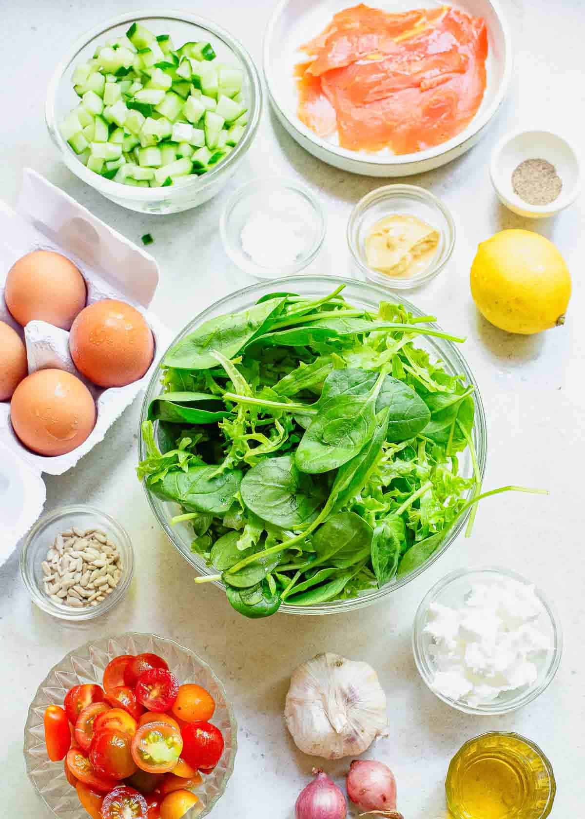 Various fresh ingredients on a table, including salmon, eggs, arugula, cucumbers, tomatoes, garlic, and lemon, arranged for meal preparation.