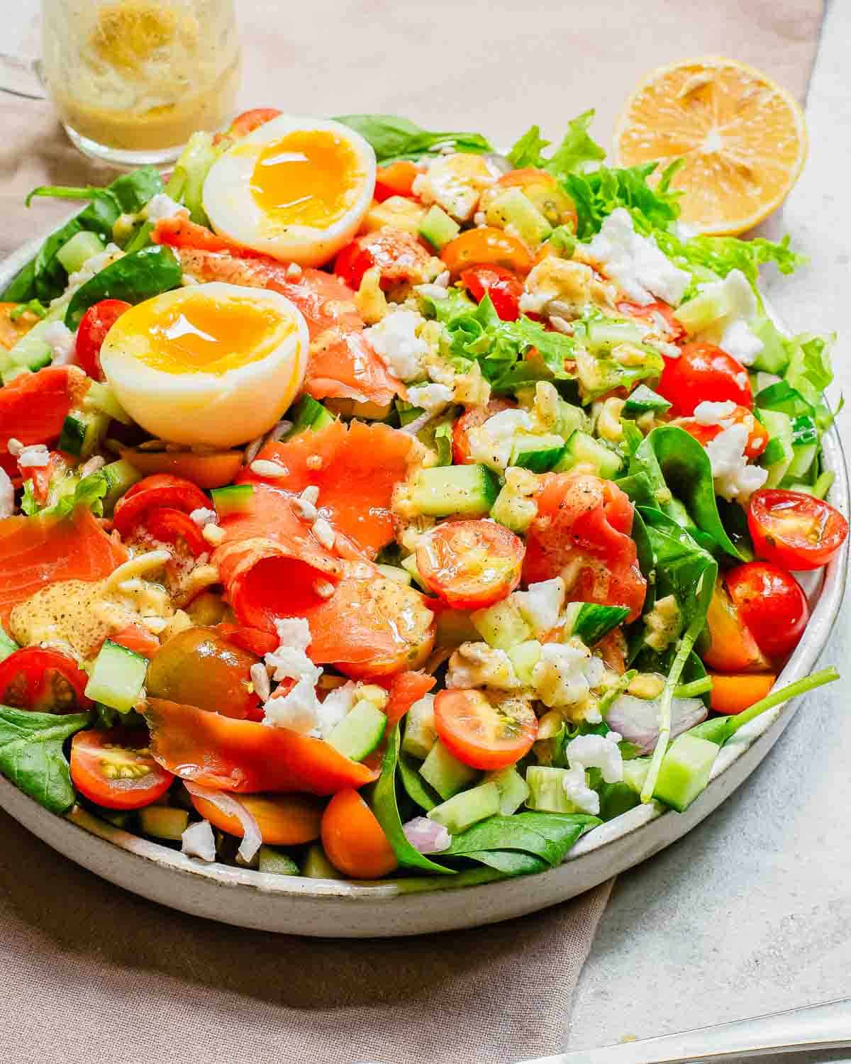 Colorful smoked salmon salad with boiled eggs, tomatoes, cucumbers, and mixed greens, served with lemon on the side.