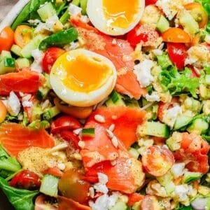 A colorful salad with smoked salmon, halved boiled egg, cucumbers, tomatoes, and mixed greens, sprinkled with seeds.