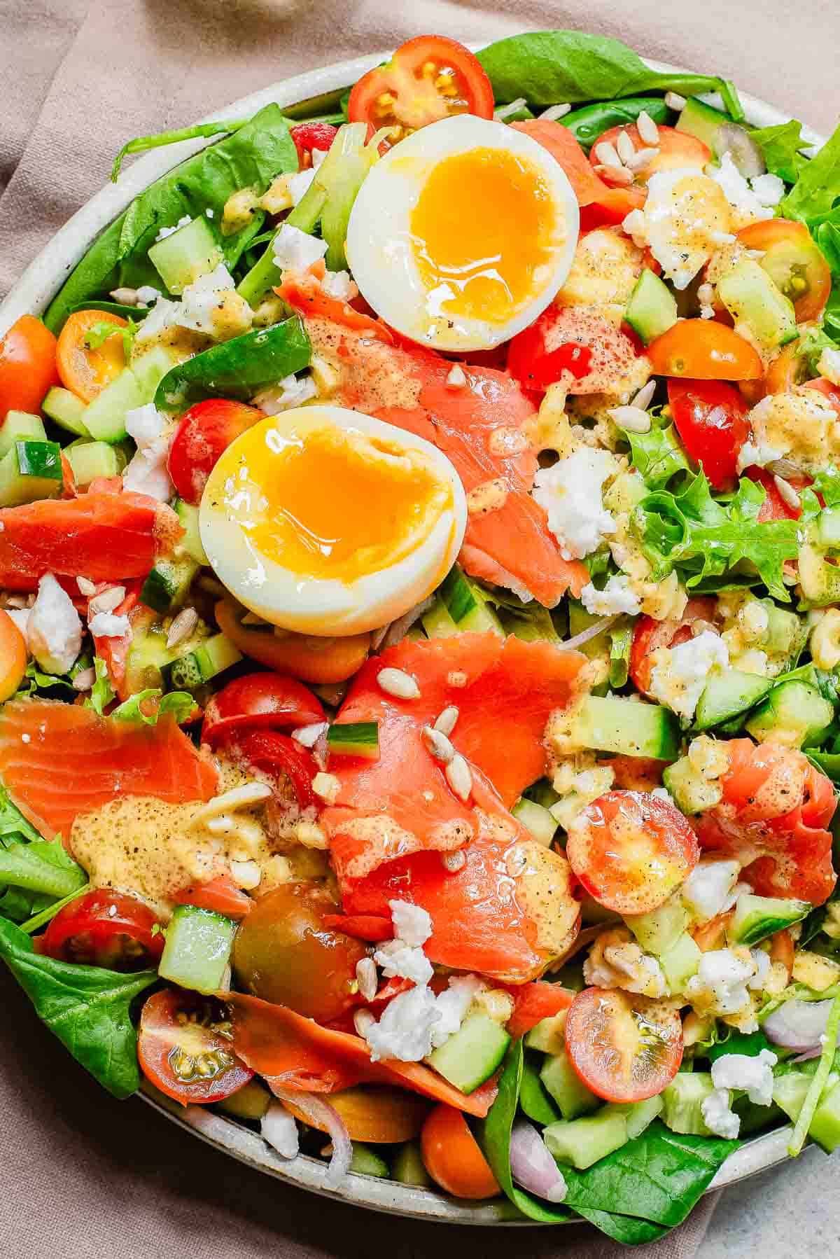 A colorful salad with smoked salmon, halved boiled egg, cucumbers, tomatoes, and mixed greens, sprinkled with seeds.