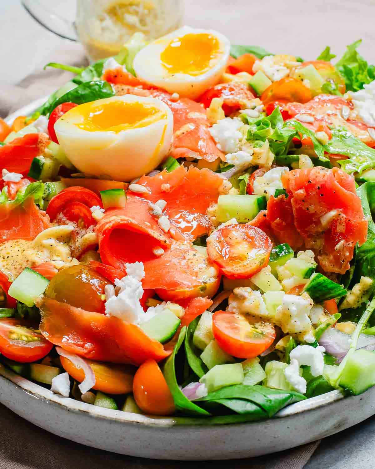 A vibrant salad with smoked salmon, boiled eggs, cherry tomatoes, cucumbers, and crumbled feta cheese, served in a gray bowl.