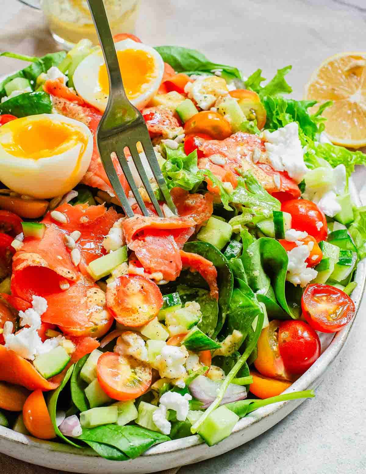 A colorful salad with smoked salmon, boiled eggs, cherry tomatoes, cucumbers, and mixed greens, topped with feta cheese and sesame seeds, served with lemon slices.