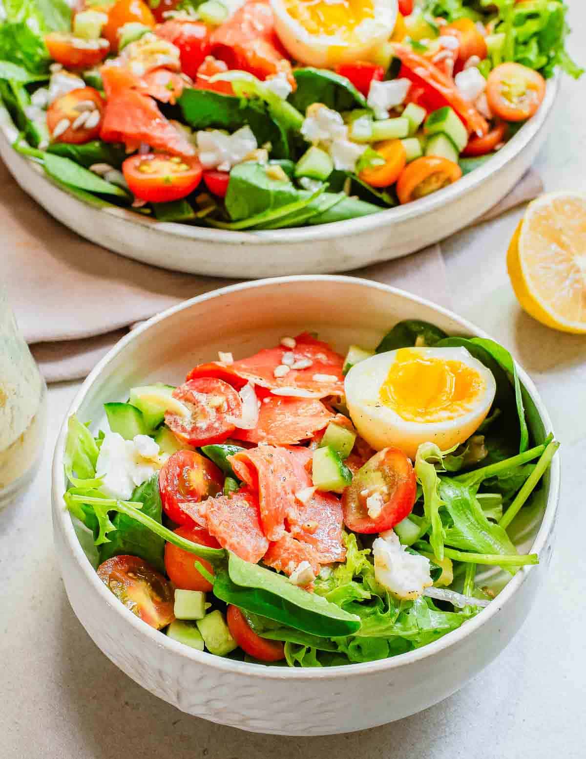 Two bowls of fresh salad with arugula, cucumbers, cherry tomatoes, smoked salmon, feta cheese, and a boiled egg, served with lemon slices on the side.