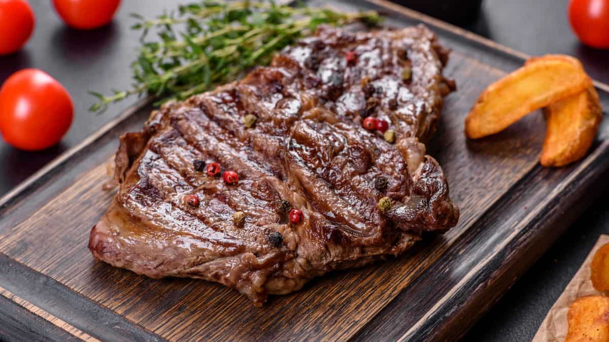 Grilled steak with peppercorns on a wooden board, accompanied by herbs, tomatoes, and potato wedges on the side.