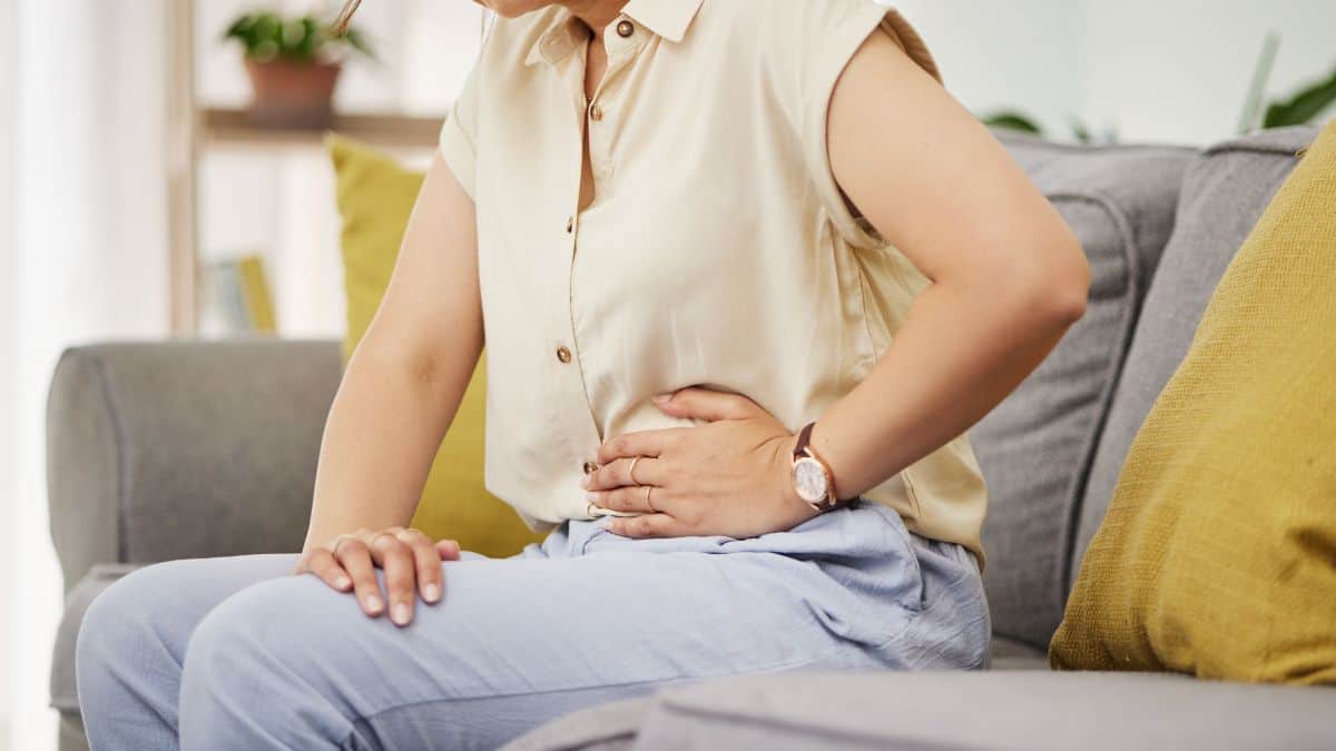 A woman sitting on a couch, holding her stomach, possibly in discomfort, wearing a beige blouse and blue pants.