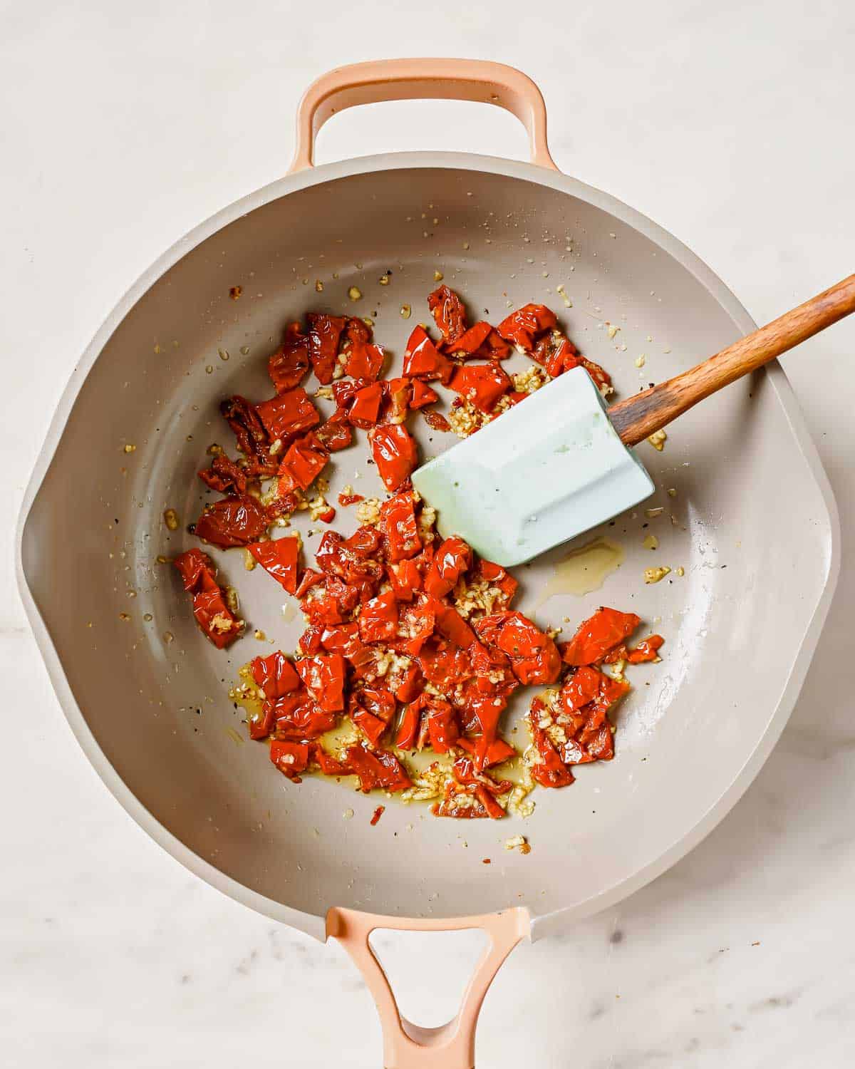 Diced sun-dried tomatoes being sautéed in a pot with a wooden spatula.