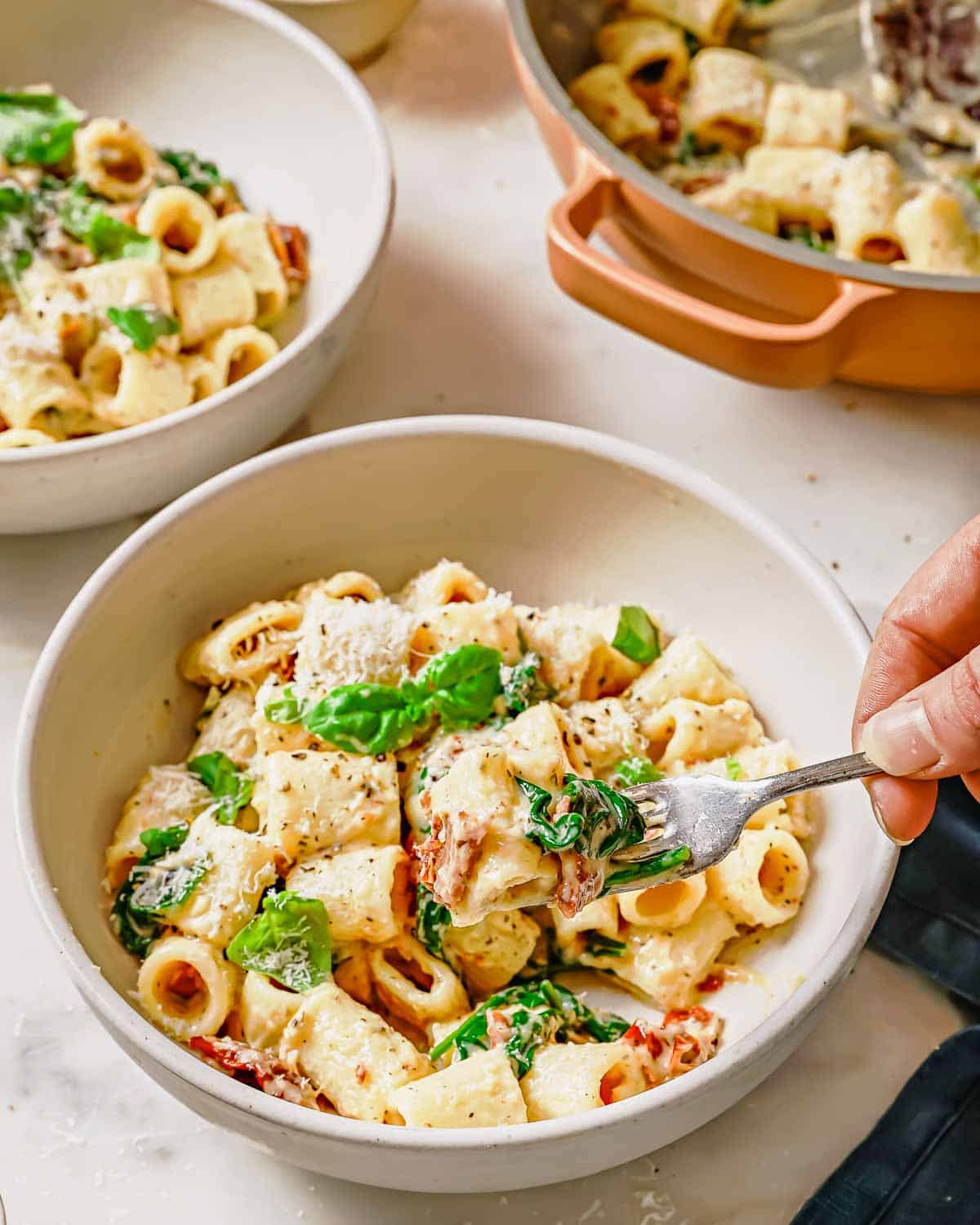 A bowl of creamy sun-dried tomato pasta garnished with herbs, being eaten with a fork.