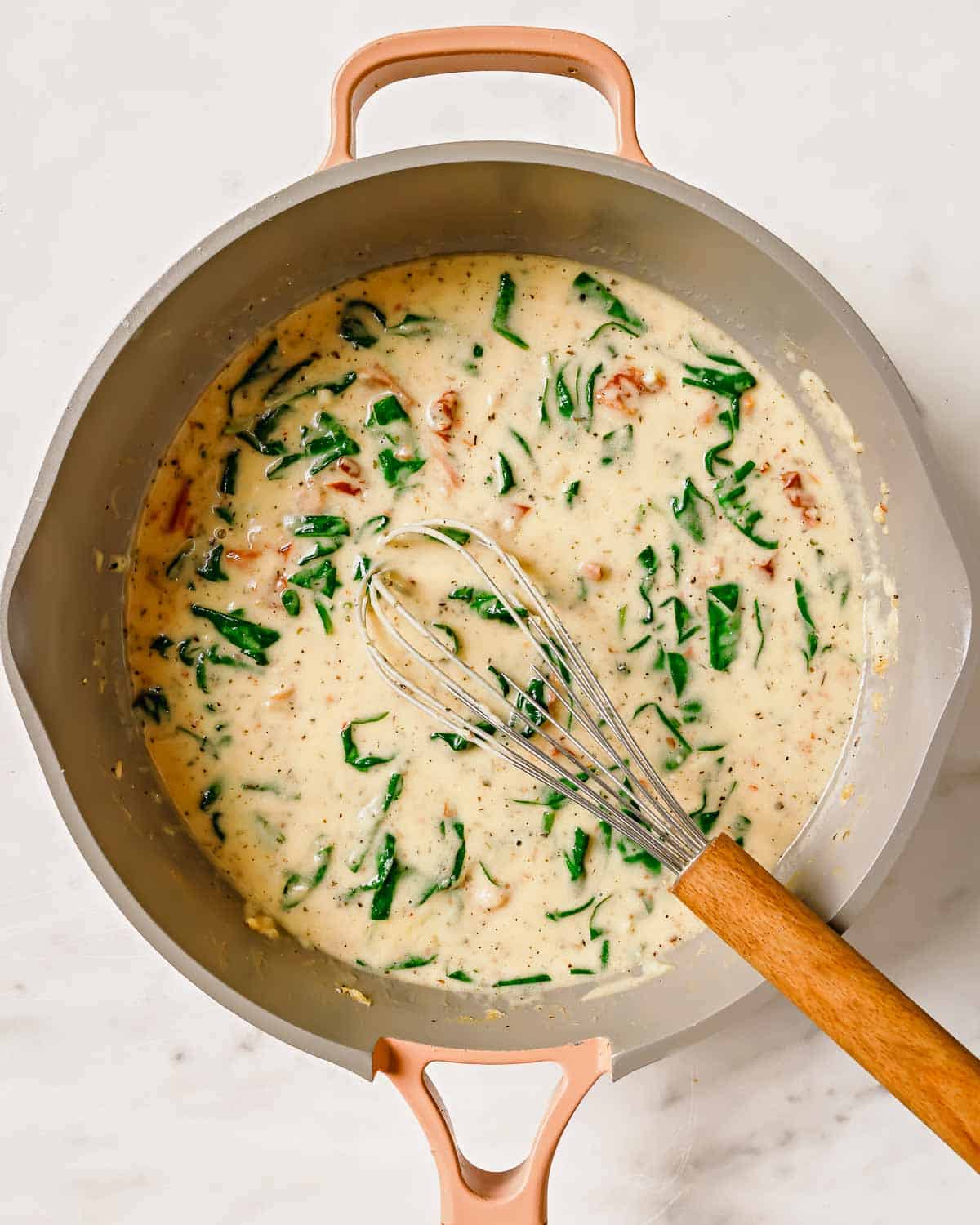 A creamy sun-dried tomato sauce with greens and spices being whisked in a pot.