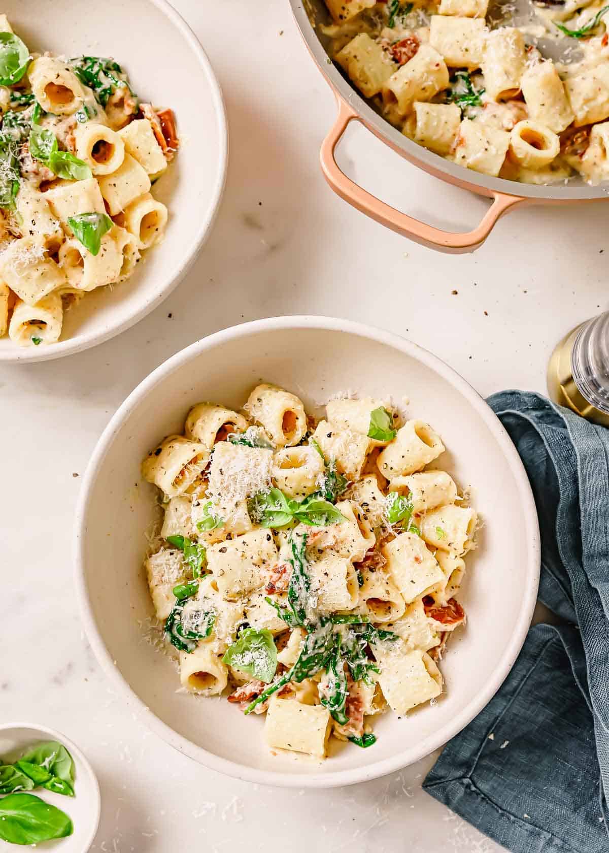 Two bowls of sun-dried tomato pasta with parmesan and herbs on a kitchen counter.