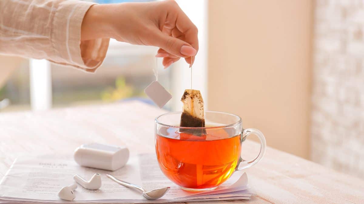 A person steeping a tea bag in a clear glass cup of hot water on a table with a newspaper and teaspoons nearby.