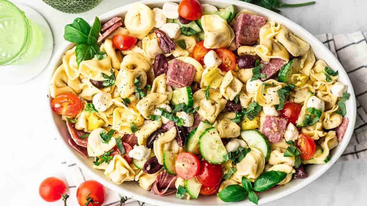 A colorful plate of pasta salad with tomatoes, cucumbers, meats, olives, and fresh herbs.