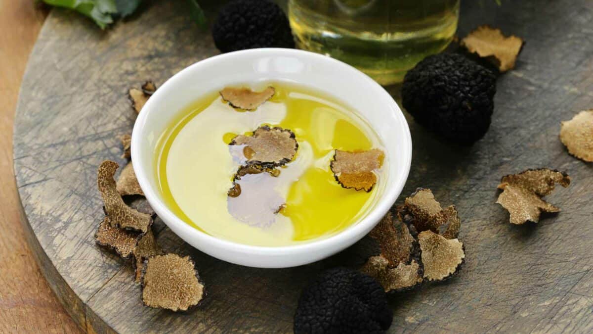 A bowl of olive oil garnished with truffle slices, surrounded by black truffles and truffle pieces on a wooden board.