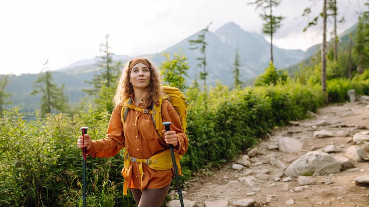 A woman in hiking gear walks on a mountain trail, carrying a backpack and trekking poles, with lush greenery and mountain peaks in the background.