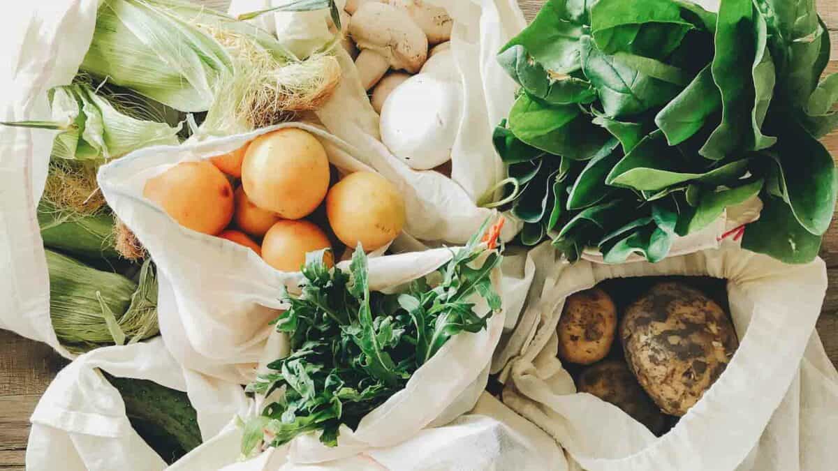 Reusable cloth bags containing fresh vegetables and fruits, including leafy greens, oranges, and potatoes, arranged on a wooden table.