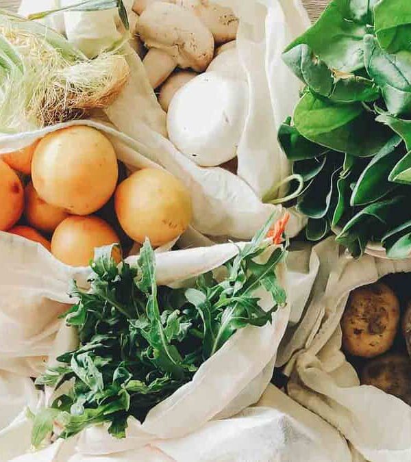 Reusable cloth bags containing fresh vegetables and fruits, including leafy greens, oranges, and potatoes, arranged on a wooden table.