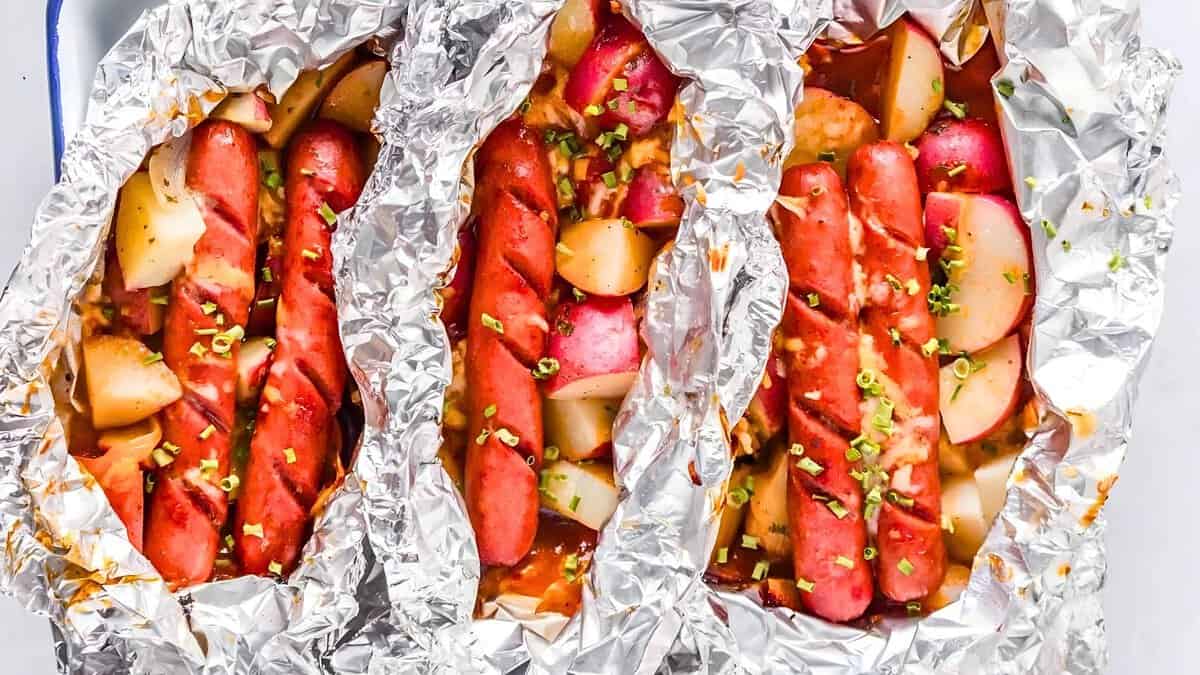 Three foil packets filled with sliced sausages, potatoes, and garnished with chopped green herbs.