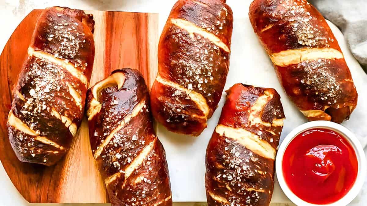 Five salted pretzel rolls on a wooden cutting board with a small bowl of ketchup on the side.