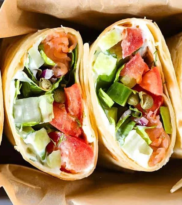 Four vegetable wraps filled with lettuce, tomatoes, cucumbers, onions, and other greens, wrapped in paper and arranged in a row.