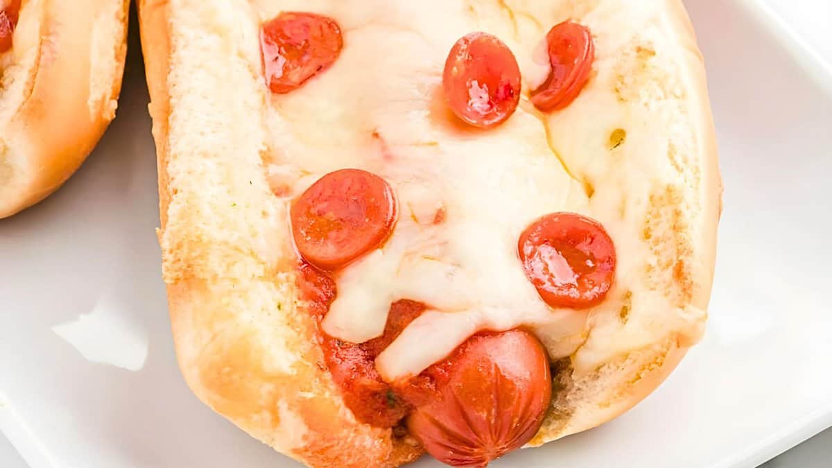 Hot dog in a bun topped with melted cheese and small pepperoni slices on a white plate.